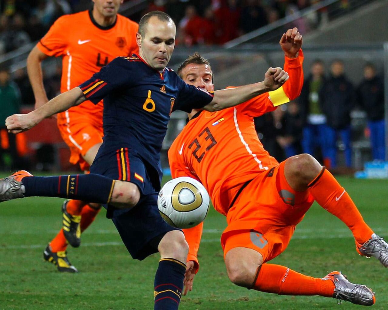 Two soccer players in a mid-match tackle during a game, one wearing a dark blue uniform, the other in bright orange, compete fiercely for the ball.