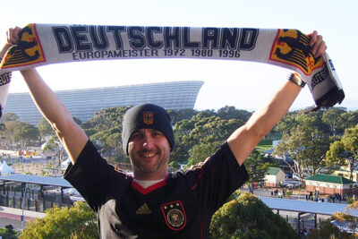 Man in a German national football team jersey joyfully holding a Deutschland scarf at a sports stadium, with trees and structures in the background.