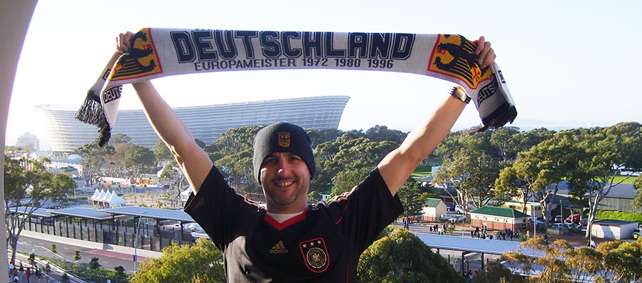 Man in a German national football team jersey joyfully holding a Deutschland scarf at a sports stadium, with trees and structures in the background.