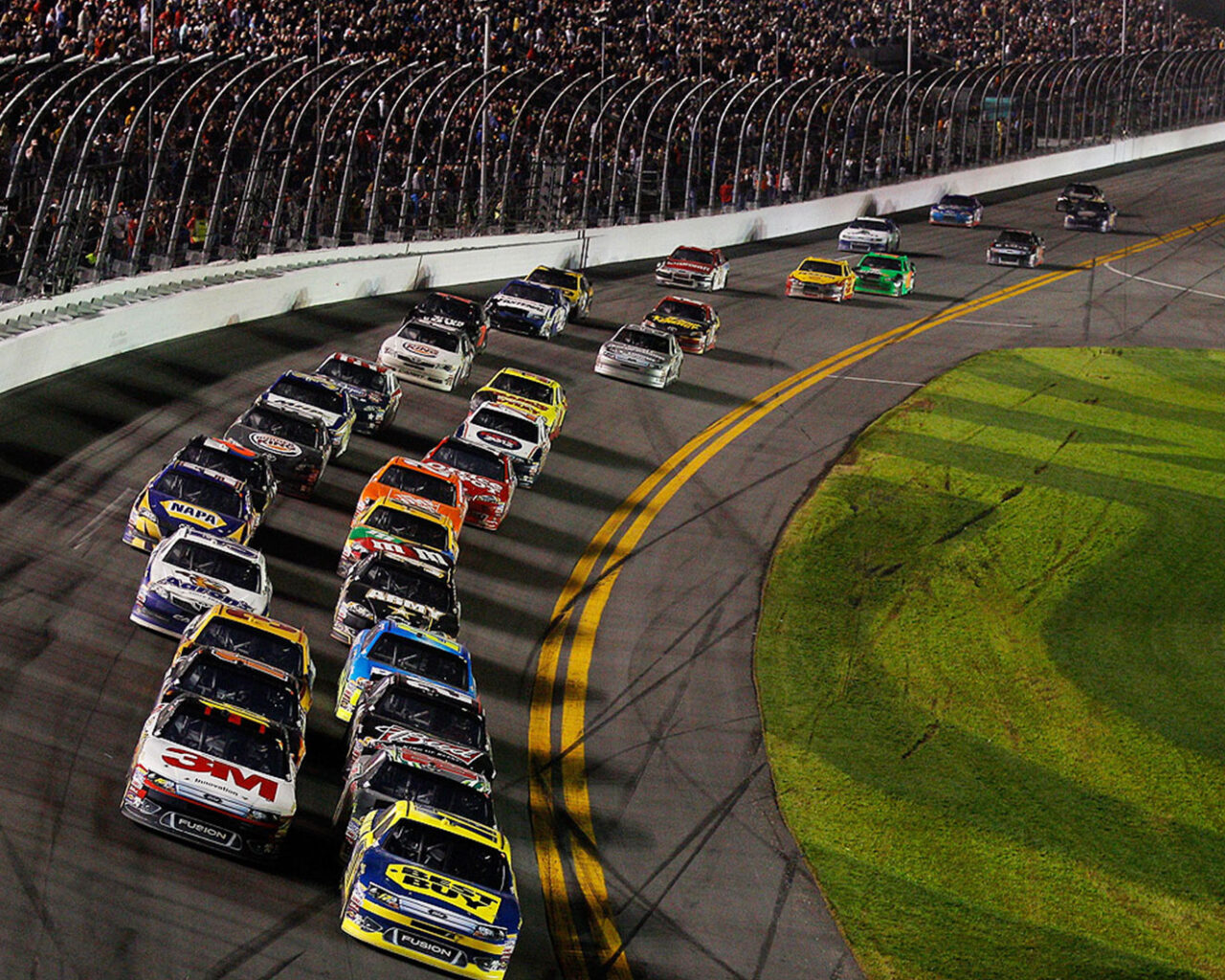 Colorful NASCAR race cars tightly packed at a curve on the Daytona 500 track during an exciting night race.