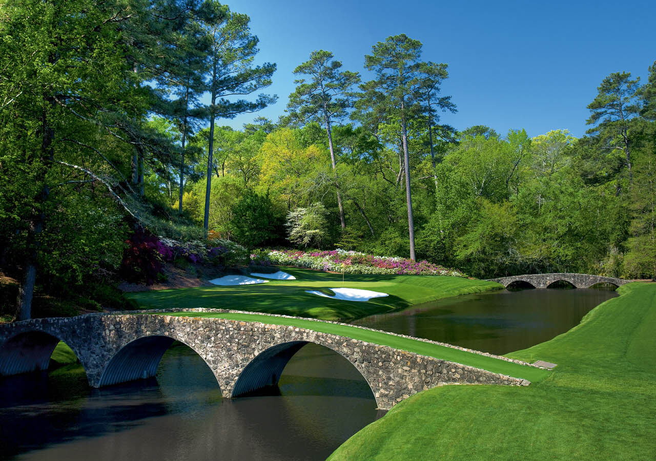 Serene view at Augusta National Golf Club featuring a stone bridge over a pond, surrounded by vibrant flowering bushes and lush trees, ideal for luxury travel guides by Roadtrips.