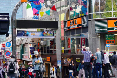 Busy street scene outside Takeshita Street in Tokyo, featuring pedestrians, colorful signage, and tourists taking photos, exemplifying vibrant urban life and cultural diversity.