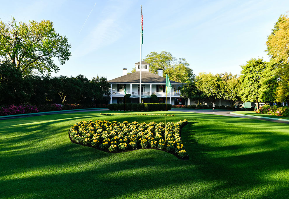Sunlight bathes the iconic clubhouse at Augusta National, framed by immaculate gardens and a floral display shaped like the United States, capturing a classic scene at The Masters.
