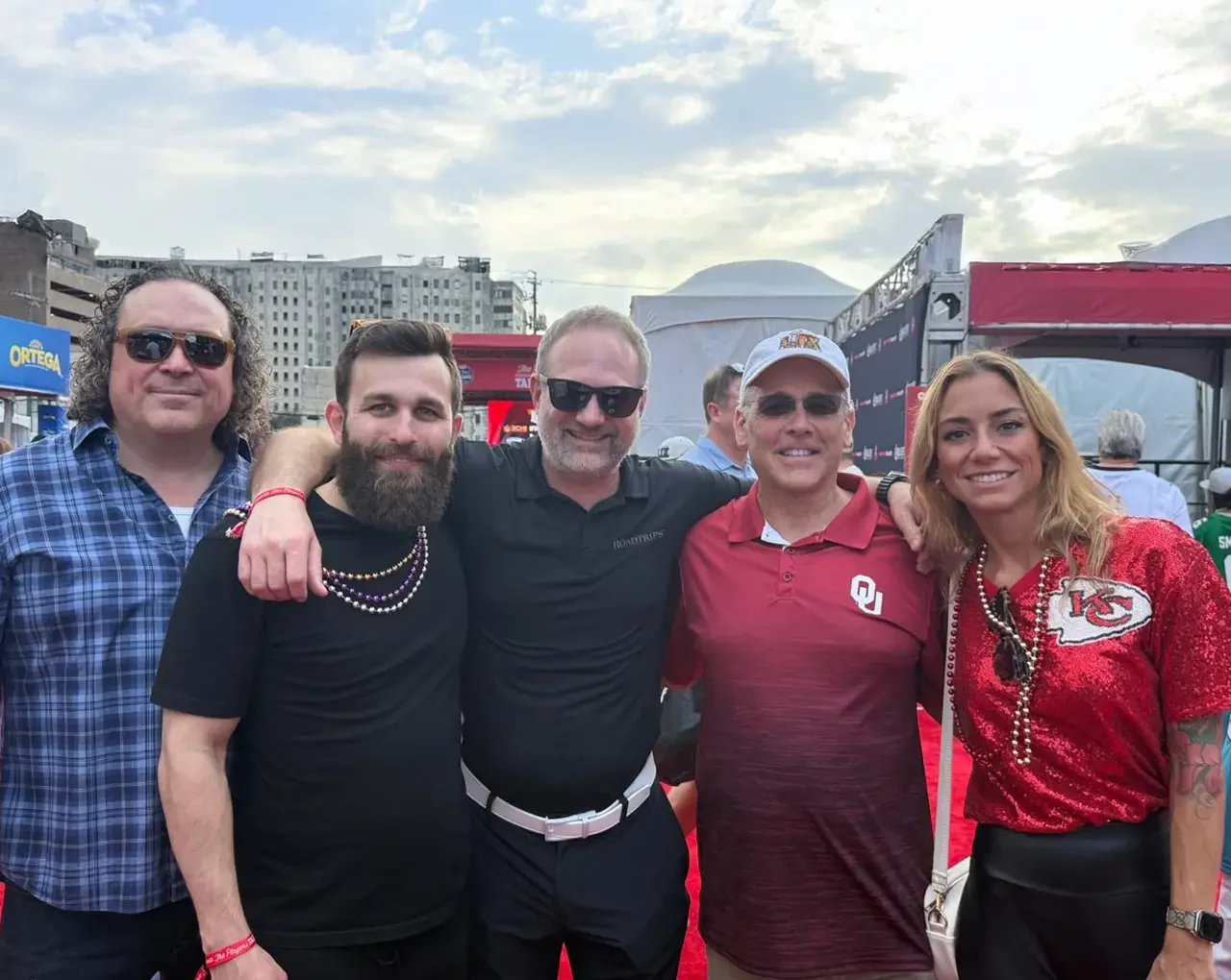 Five adults smiling at a tailgate party, with a woman in a glittering red jacket and men in casual attire, hinting at an exciting NFL event.