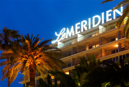 A twilight view of the Le Meridien hotel, prominently featuring its illuminated sign, flanked by palm trees.