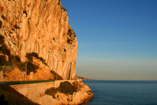 Sunset view of a coastal road beside a towering cliff over the Mediterranean, reflecting the beauty of Monte Carlo, a prime location for the Monaco Grand Prix.