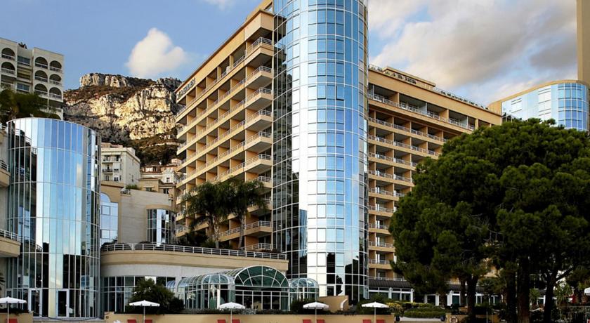 Modern hotel with reflective blue glass towers and balconies overlooks a rocky hill, located near Monte Carlo, ideal for Monaco Grand Prix attendees.