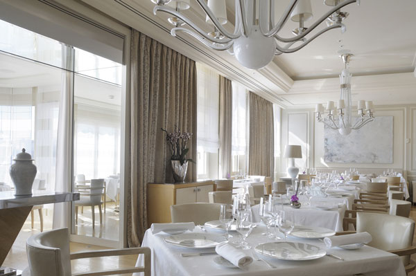 Elegant dining room in a luxury hotel featuring white tablecloths, crystal chandeliers, and modern art, located near the Monaco Grand Prix accommodations in Monte Carlo.