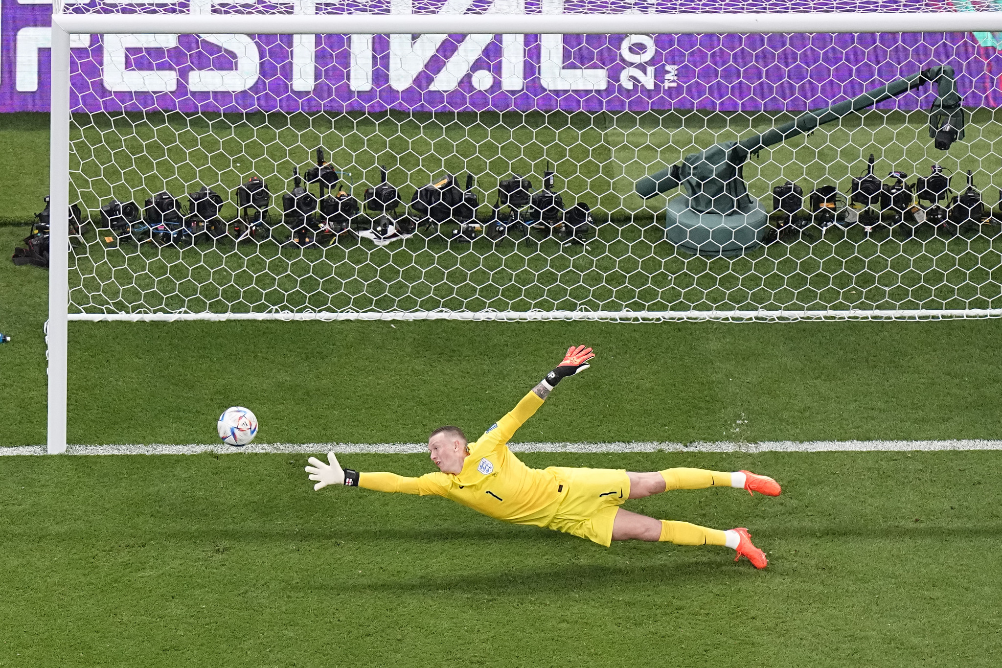 Soccer goalkeeper in yellow dives towards the ball during a match, with a view of the goal and photographers in the background, relevant for discussions about luxury travel packages to major sports events.