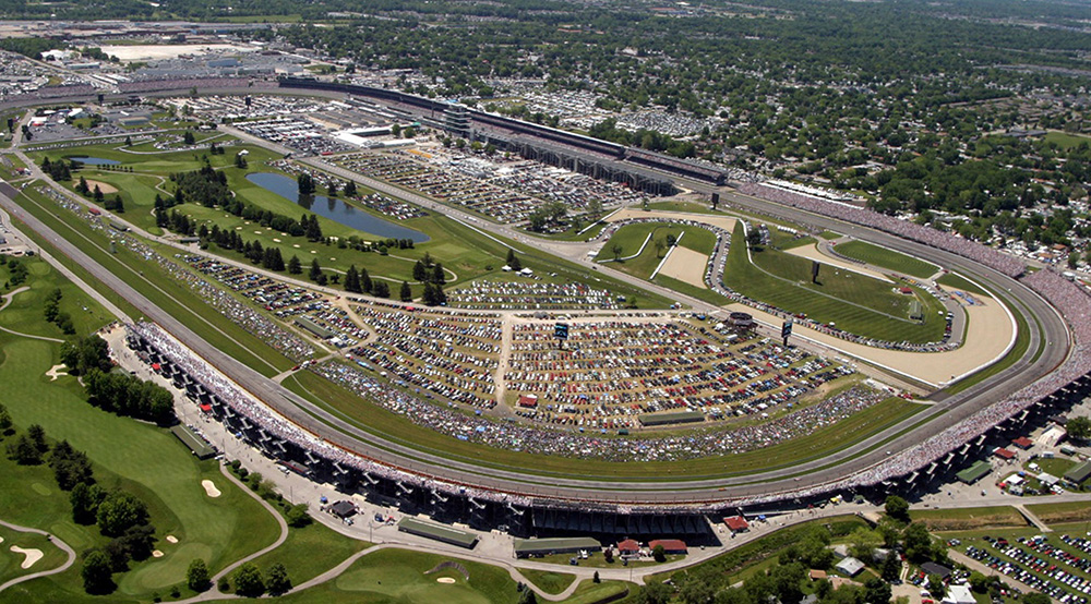 Aerial view of the Indianapolis Motor Speedway filled with spectators and parked cars during a major racing event, perfect for first-timers exploring luxury travel guides by Roadtrips.