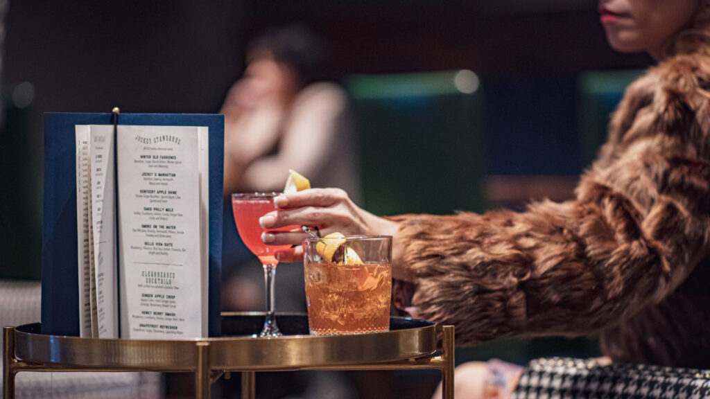 At a luxury hotel, a person in a fur coat enjoys a cocktail next to a menu, capturing the hospitality near Churchill Downs, ideal for Kentucky Derby visitors in Louisville.