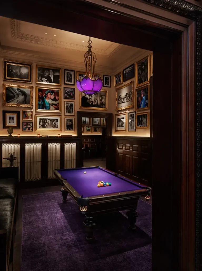 View into a sophisticated billiard room at The New York EDITION, featuring a purple pool table, ornate purple chandelier, and walls adorned with framed photographs. Ideal for guests seeking luxury accommodations near Ryder Cup venues.