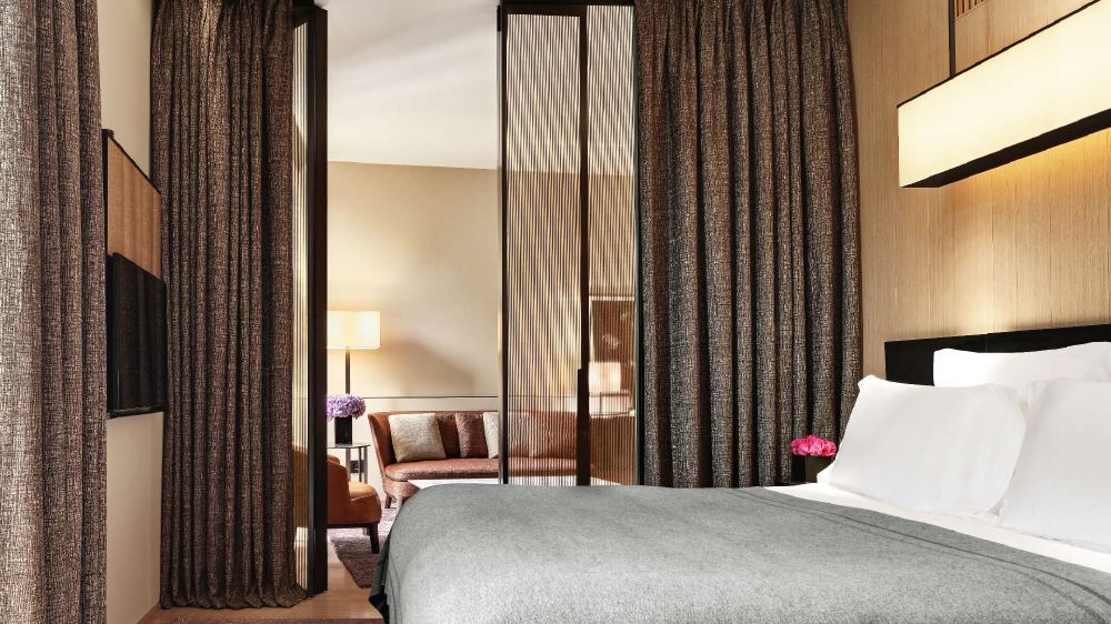 Luxurious hotel room at Bvlgari Hotel Milano, featuring a large bed with white linens, upholstered seating, and elegant, floor-to-ceiling drapery, ideal for guests visiting the Italian Grand Prix in Milan.