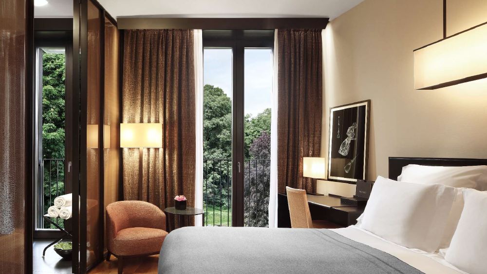 A well-appointed hotel room in Milan features an open balcony door revealing lush greenery, elegant interior design, and a cozy atmosphere, suited for visitors to the Italian Grand Prix.