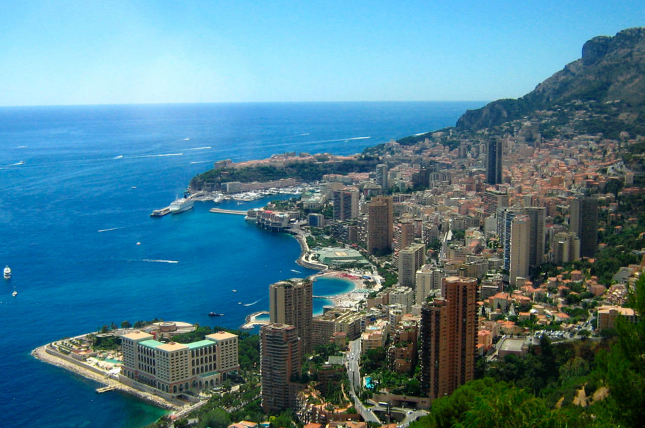 Aerial view of Monaco's dense urban layout, featuring high-rise buildings along the coastline with a backdrop of mountains, highlighting a luxury travel destination near the Monaco Grand Prix.