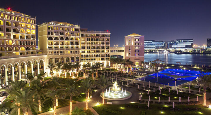 Luxurious hotel in Abu Dhabi, UAE, illuminated at night with a view of palm trees, a fountain, and lit pools, overlooking the sparkling cityscape. Ideal for guests attending Abu Dhabi Grand Prix events.