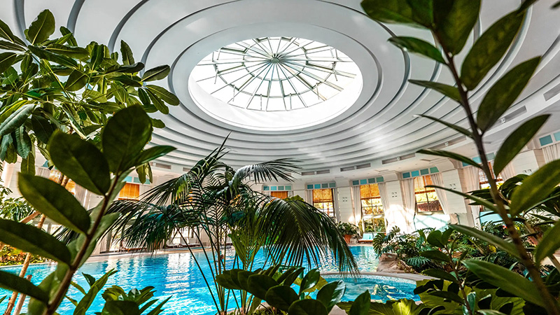Lush green plants surround the vibrant indoor pool at the Monte Carlo Bay Hotel, Monaco, beneath a large skylight, offering a luxurious accommodation option for Monaco Grand Prix attendees.