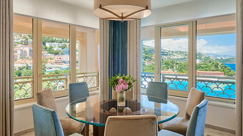 Interior view of a luxurious dining area with a round glass table and plush chairs, overlooking the scenic hills and ocean in Monaco, near the Monte Carlo Bay Hotel.