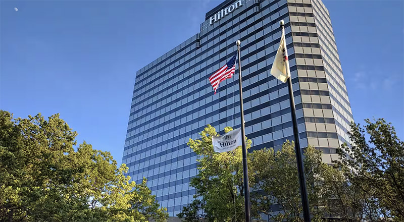 Hilton hotel in New York, showing the building's exterior under a clear sky with an American flag and Hilton flag prominently displayed. Ideal for upcoming World Cup hotel accommodations.