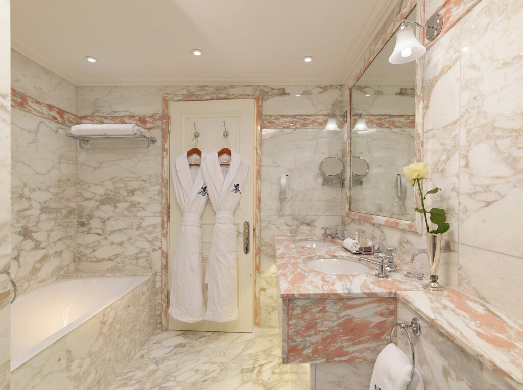 An elegant marble bathroom at The Ritz London, featuring a bathtub, mirrored walls, and robes, ideal for visitors attending the Wimbledon Championships.