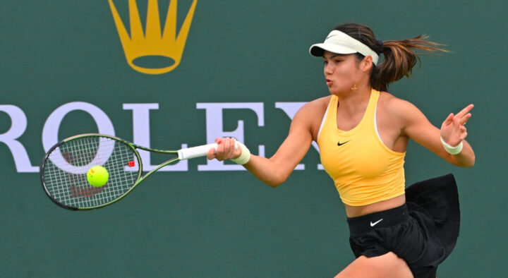 A female tennis player in a yellow top and black skirt reaches to hit a tennis ball at a court.