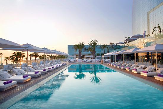 Sunset view of W Hotel's outdoor pool in Barcelona, featuring aligned sun loungers and palm trees reflecting in the tranquil water, ideal for guests attending the Spanish Grand Prix.