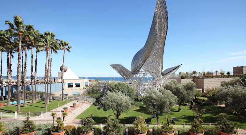 Palm trees and lush greenery surround a large, metallic fish sculpture near the seafront, showcasing a popular locale potentially linked to the Spanish Grand Prix event offerings.