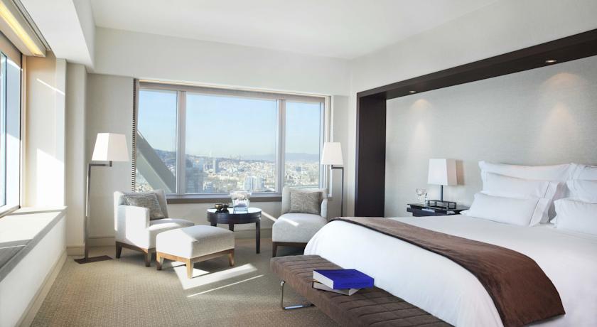 Luxurious hotel room with a panoramic view of Barcelona, featuring a large bed, armchairs, and a clear day outside—ideal for Spanish Grand Prix accommodations.