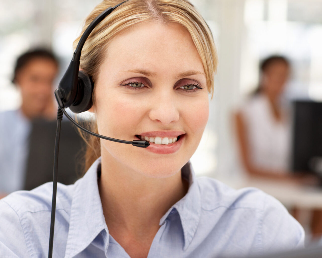 A customer service representative wearing a headset smiles while working in a busy office environment.