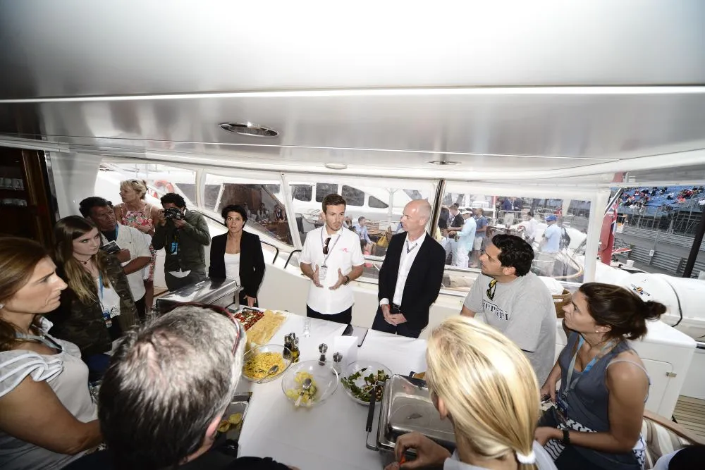 Chef demonstrates cooking to an attentive group on a yacht, providing an exclusive group hospitality experience, emphasizing global and trusted services with over 30 years of expertise.